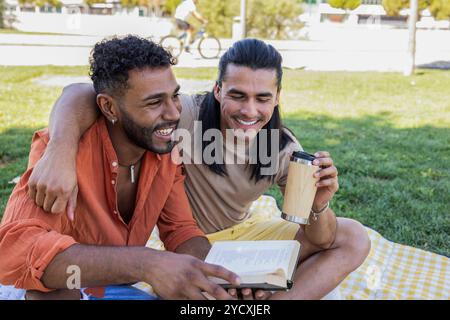 Couple gay, représentant un mélange d'ethnies latino-américaine et afro-américaine, se détendre dans un parc ; l'un sirote un verre tandis que l'autre lit un livre, Ill Banque D'Images