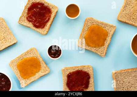 Vue aérienne de tranches de pain multigrains avec des quantités variables de confiture de fraises et de pêches étalées, présentées soigneusement sur un fond bleu subtil Banque D'Images
