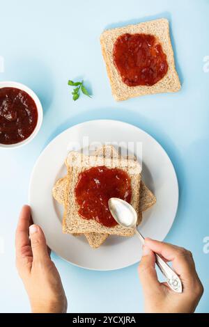 Vue aérienne des mains d'une personne étalant de la confiture de fraises sur des toasts à côté d'un bol de confiture et une feuille de menthe sur un fond bleu Banque D'Images