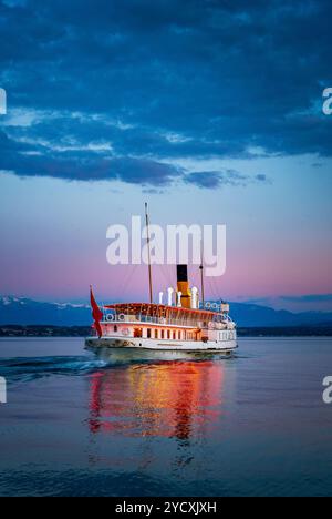 Un bateau à vapeur à aubes classique navigue sur le lac Léman, le lac Léman près de Coppet, en Suisse, contre un ciel de coucher de soleil saisissant avec des silhouettes alpines dans le Banque D'Images