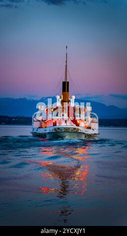 Un élégant bateau à aubes vintage navigue sur le lac Léman, le lac Léman près de Coppet, en Suisse, ses lumières se reflétant sur l'eau au crépuscule contre un Banque D'Images