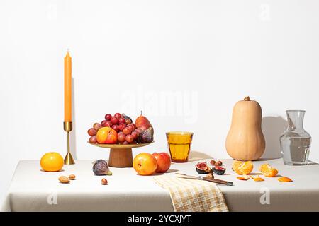 Une nature morte présentant des fruits assortis comme des raisins, des pêches et des oranges disposés sur un bol de fruits en bois, accompagné d'une grande bougie, un butternut s Banque D'Images
