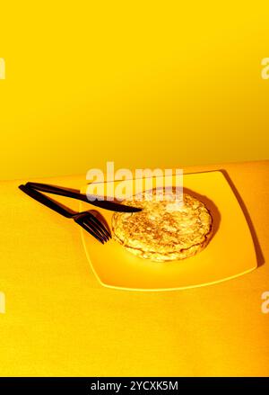 Une tortilla espagnole fraîchement cuite se trouve sur une assiette jaune vif, complétée par un fond jaune et des ombres douces, avec des ustensiles noirs Banque D'Images