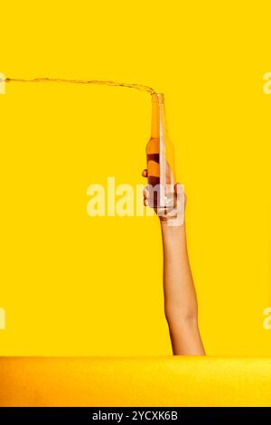 Une image créative représentant une main tenant une bouteille en verre d'où une bière claire jaillit, le tout sur un fond jaune vif Banque D'Images