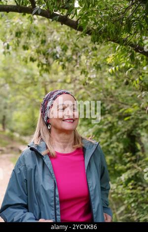 Une femme mature portant un foulard coloré et une tenue sportive poutres avec joie tout en faisant du jogging à travers un parc verdoyant et luxuriant, son expression reflète le contentm Banque D'Images