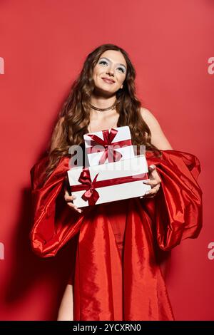 Une jeune femme sourit avec éclat alors qu'elle tient des cadeaux emballés, rayonnant de joie festive dans un décor rouge. Banque D'Images