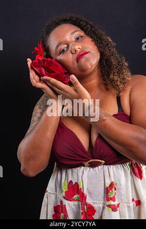 Belle femme noire portant des vêtements traditionnels, tenant une rose rouge à côté de son visage et posant. Isolé sur fond noir. Photo studio. Banque D'Images