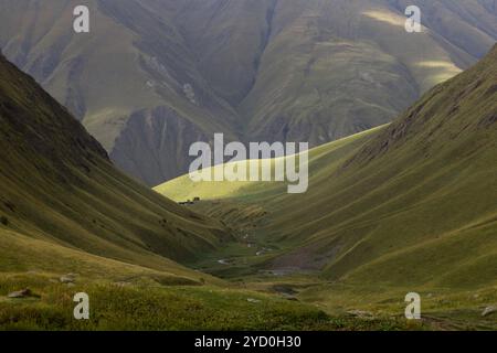 une vallée tranquille entourée de collines escarpées et vallonnées et de hautes montagnes au loin. Le paysage est couvert d'herbe verte luxuriante, avec la lumière du soleil b Banque D'Images