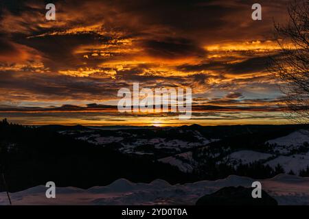 Alors que le soleil se couche sous l'horizon, des teintes orange et jaunes vibrantes remplissent le ciel au-dessus de paisibles montagnes enneigées, créant une veille hivernale pittoresque Banque D'Images