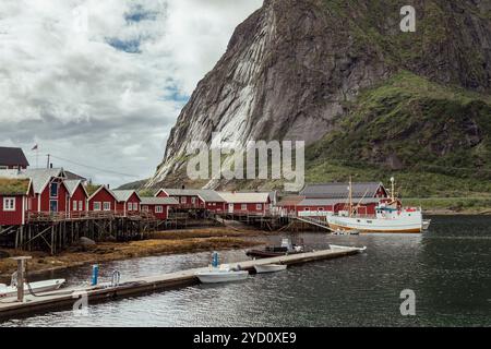 Un port pittoresque dans les îles Lofoten présente des maisons rouges vibrantes et des bateaux de pêche, nichés sur des paysages montagneux spectaculaires sous un ciel nuageux Banque D'Images