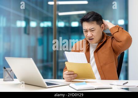 Homme d'affaires asiatique semble surpris de recevoir le courrier au bureau. L'expression d'une préoccupation indique une mauvaise nouvelle ou inattendue. Courrier enveloppé tenu dans la main, ouvrez l'ordinateur portable sur le bureau. Banque D'Images