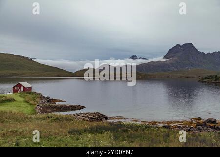 Un lac tranquille reflète les hautes terres et les montagnes environnantes des îles Lofoten, en Norvège. Une petite cabane rouge ajoute du charme au paysage pittoresque UDE Banque D'Images