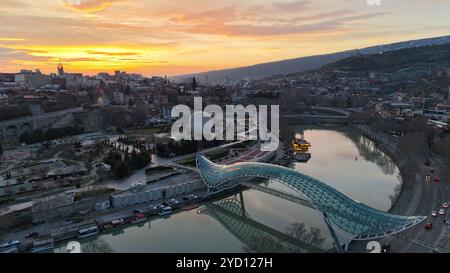 L'emblématique Pont de la paix enjambe la rivière Kura au coucher du soleil à Tbilissi, Géorgie, sa structure en verre et en acier illuminée contre un ciel doré. Banque D'Images