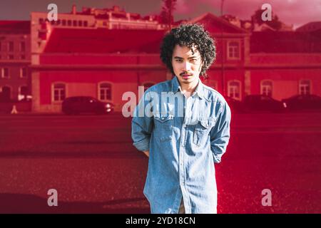 Portrait d'un jeune mannequin philippin aux cheveux bouclés, portant une chemise en denim bleu clair, debout devant un reflet urbain rouge de Lisbonne, Port Banque D'Images