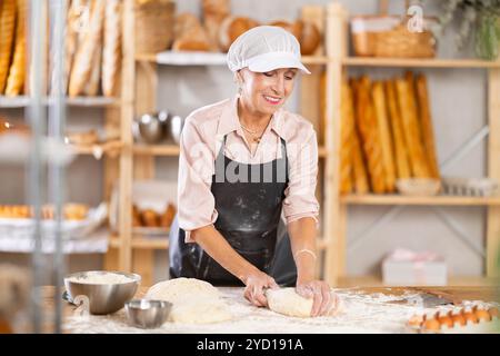 Femme chef mûre se tient sur son banc de travail, pétrissant et façonnant la pâte pour faire du pain, des croissants et des baguettes Banque D'Images