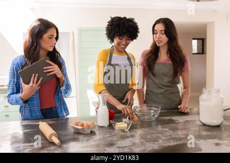 Temps de Noël, amies féminines multiraciales faisant cuire, en utilisant la tablette pour des conseils de recette, à la maison Banque D'Images