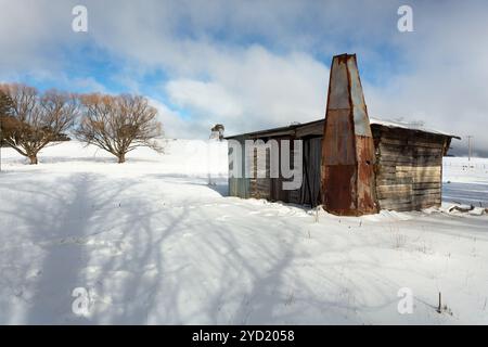 Ancienne cabane en bois, cabane ou écurie dans un enclos rural avec couverture complète des chutes de neige en hiver. Le ciel bleu fait une brève apparition entre les fluries de neige Banque D'Images