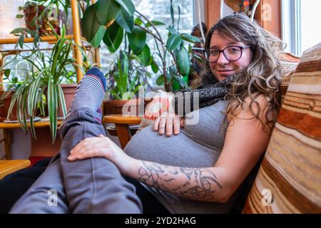 Heureux et détendu expectant parents s'asseoir ensemble dans la salle de séjour, la perspective de la première personne que l'homme repose jambes dans le tour de la femme fortement enceinte Banque D'Images