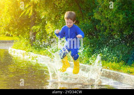 Un garçon heureux en bottes de caoutchouc saute dans les flaques d'eau. Le garçon saute dans une flaque . Mauvais temps. flaques d'eau après la pluie. Un enfant en bouc de caoutchouc Banque D'Images