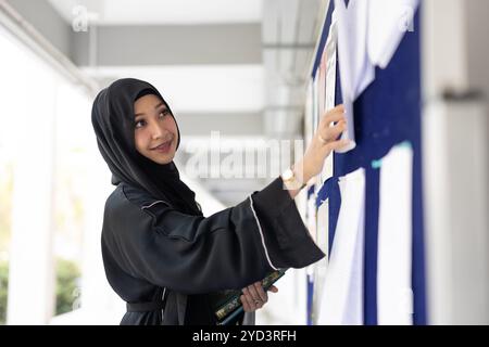 Étudiant musulman à l'université souriant vêtu de niqab noir trouvant du travail en regardant le panneau d'affichage. Dame chador noire arabe. Banque D'Images