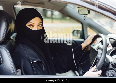 Femme du moyen-Orient conduisant un habillage de niqab noir couvert de visage de voiture, regardant caméra Banque D'Images