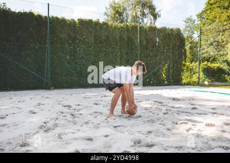 Joueur de volley-ball ramassant la balle sur un terrain de sable, se préparant pour le prochain match dans un match de volley-ball de plage occasionnel. Capturé dans un extérieur calme Banque D'Images