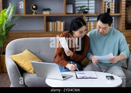 Couple asiatique examinant des documents financiers dans le salon, illustrant le travail d'équipe, la gestion des finances à domicile. Homme et femme utilisant un ordinateur portable, téléphone et paperasse pour la planification, la communication. Banque D'Images