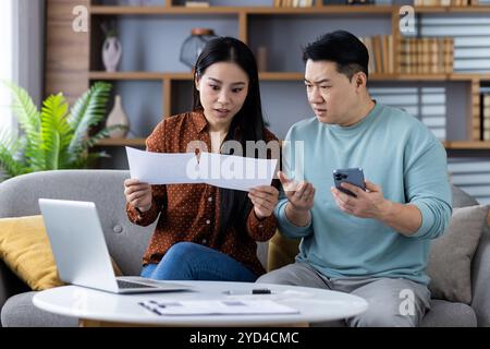Couple asiatique analysant des papiers financiers avec ordinateur portable et smartphone. L'homme et la femme expriment leur inquiétude tout en abordant les questions de budget familial. Concept de finance familiale, de budgétisation et d'intégration technologique Banque D'Images