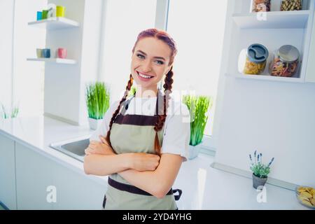 Photo d'adorable fille mignonne portant tablier bras croisés cuisine maison maison lumière de jour à l'intérieur Banque D'Images