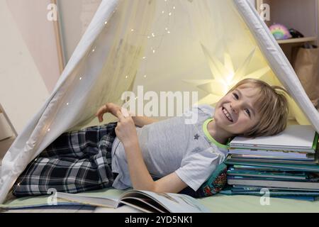 Mignon garçon de 7 ans se trouve avec sa tête sur des piles de livres dans la tente de jeu des enfants. Petit amoureux de la lecture de livres, fantasmes d'enfants, ch. intéressant Banque D'Images