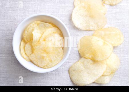 Chips de pommes de terre salées dans un bol blanc sur tissu de lin. Chips fraîches et prêtes à manger, fines tranches de pommes de terre frites. Banque D'Images