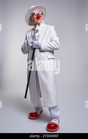 Un clown âgé dans un manteau blanc et un chapeau avec un bâton de marche regardant vers le haut. L'homme porte de grandes bottes de clown en cuir verni rouge. Banque D'Images