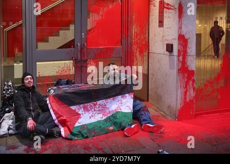 Londres, Angleterre, Royaume-Uni. 25 octobre 2024. Des activistes de Palestine action brandissent un drapeau palestinien pour dissimuler la valise renforcée qui contient leurs mains enchaînées dans la porte des bureaux de l'APCO. Les activistes bloquent les entrées des bureaux d'APCO dans le Strand pour la deuxième fois en un mois à Londres, s'attachant à un verrou de valise, après avoir pulvérisé l'extérieur du bâtiment avec de la peinture rouge sang. APCO est embauché par Elbit Systems pour faire pression sur les politiciens en leur nom. Palestine Actionists continuent de prendre des mesures directes contre les entreprises qui permettent à la compagnie israélienne d'armement Elb Banque D'Images