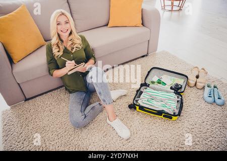 Photo d'adorable belle femme joyeuse assise sur le sol écrivant des plans d'organisation des vêtements dans valise appartement à l'intérieur plat Banque D'Images