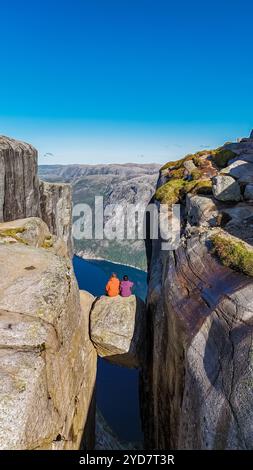 Deux individus sont assis sur un rebord rocheux étroit surplombant une vallée profonde dans la falaise NorwaynKjeragbolten Banque D'Images