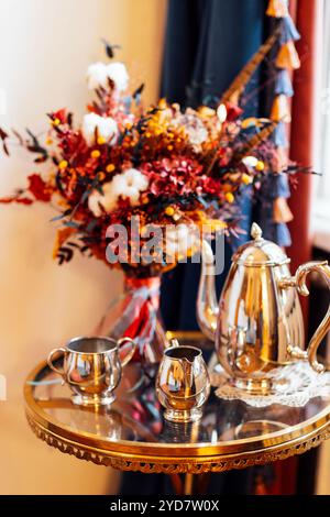 Une belle table basse ronde en verre avec une bouilloire en métal, un pot à lait et une tasse. Bouquet d'automne de fleurs avec coton et elegan Banque D'Images