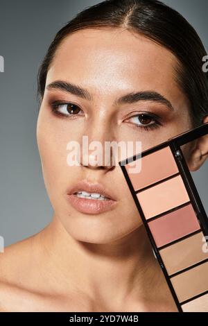 Une belle femme met en valeur ses traits tout en tenant une palette de maquillage dans un environnement élégant. Banque D'Images