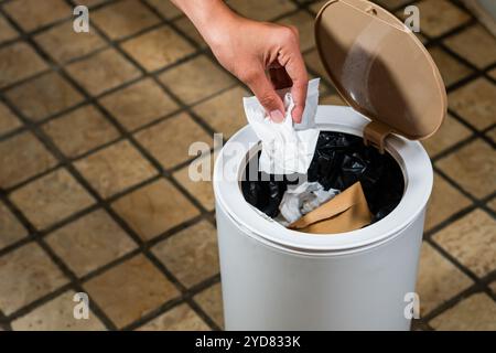 Une main laissant tomber un morceau de papier froissé dans une poubelle blanche avec un couvercle. La poubelle est partiellement remplie de divers types de déchets, et le fl Banque D'Images