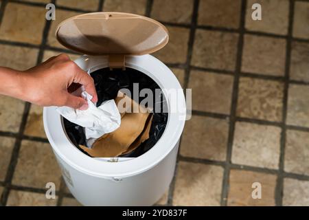 Une main laissant tomber un morceau de papier froissé dans une poubelle blanche avec un couvercle. La poubelle est partiellement remplie de divers types de déchets, et le fl Banque D'Images