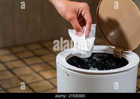 Une main laissant tomber un morceau de papier froissé dans une poubelle blanche avec un couvercle. La poubelle est partiellement remplie de divers types de déchets, et le fl Banque D'Images