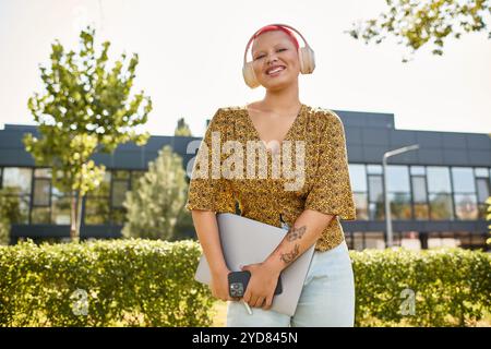 Une femme chauve avec un sourire chaleureux tient son ordinateur portable sous le soleil brillant, profitant de sa journée de travail à l'extérieur. Banque D'Images