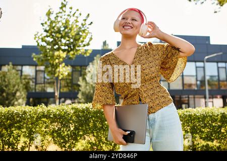 Une joyeuse femme chauve écoute de la musique avec des écouteurs, tenant son ordinateur portable tout en souriant dehors. Banque D'Images