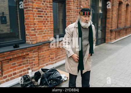 Portrait d'homme âgé sans-abri en manteau beige et foulard vert debout dans la rue à côté de maigres possessions, confronté à la misère et à la misère Banque D'Images