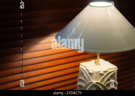 Un gros plan d'une lampe élégante avec une base texturée et un abat-jour conique, éclairée doucement sur une toile de fond de stores en bois. Banque D'Images
