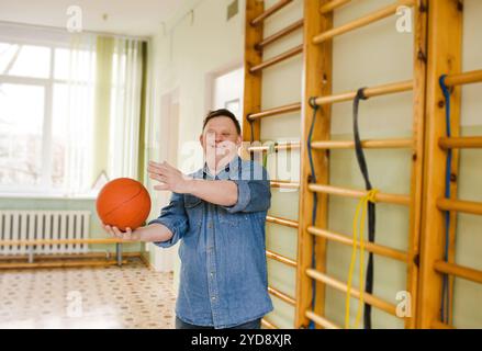 Un jeune homme atteint du syndrome de Down joue au basket-ball dans une salle de gym. Il sourit avec éclat, démontrant la joie et les bienfaits de l’activité physique pour la veille Banque D'Images