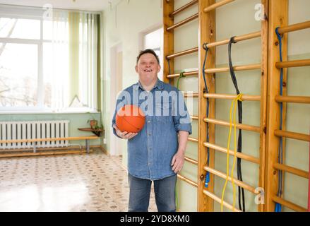 Un jeune homme atteint du syndrome de Down se tient dans un gymnase, tenant un ballon de basket et souriant avec éclat. Il respire la confiance et la joie, démontrant le pouvoir Banque D'Images