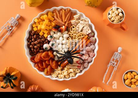 Panneau de charcuterie sucrée d'Halloween avec citrouille, pop-corn, fruits secs, bonbons au chocolat de couleur, araignée, craquelins sur fond orange. Nourriture de fête pour Banque D'Images