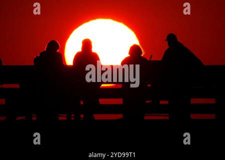 Île de Palms, États-Unis. 25 octobre 2024. Les touristes silhouettés par le lever du soleil regardent le soleil orange géant se lever sur l'océan Atlantique depuis l'embarcadère de l'île de Palms sur Front Beach, le 25 octobre 2024 à l'île de Palms, Caroline du Sud. Un temps ensoleillé et chaud est prévu pour le pays bas pour les prochaines semaines. Crédit : Richard Ellis/Richard Ellis/Alamy Live News Banque D'Images