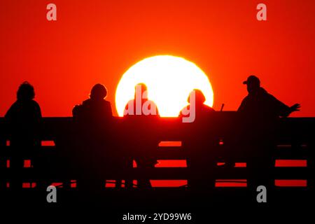 Île de Palms, États-Unis. 25 octobre 2024. Les touristes silhouettés par le lever du soleil regardent le soleil orange géant se lever sur l'océan Atlantique depuis l'embarcadère de l'île de Palms sur Front Beach, le 25 octobre 2024 à l'île de Palms, Caroline du Sud. Un temps ensoleillé et chaud est prévu pour le pays bas pour les prochaines semaines. Crédit : Richard Ellis/Richard Ellis/Alamy Live News Banque D'Images