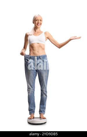 Jeune femme pesant sur une balance et essayant sur une paire de jeans isolé sur fond blanc Banque D'Images